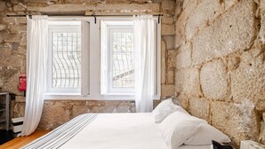 A bedroom with a stone wall, a bed with white bedding, a window with white curtains, and a radiator. - Vitória, The Studio in the Heart of Porto World Heritage (Porto)