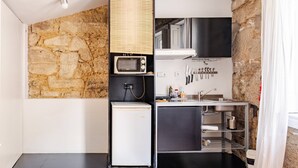 A compact kitchen with a stone wall, a microwave, a small refrigerator, and a sink with hanging utensils. - Vitória, The Studio in the Heart of Porto World Heritage (Porto)