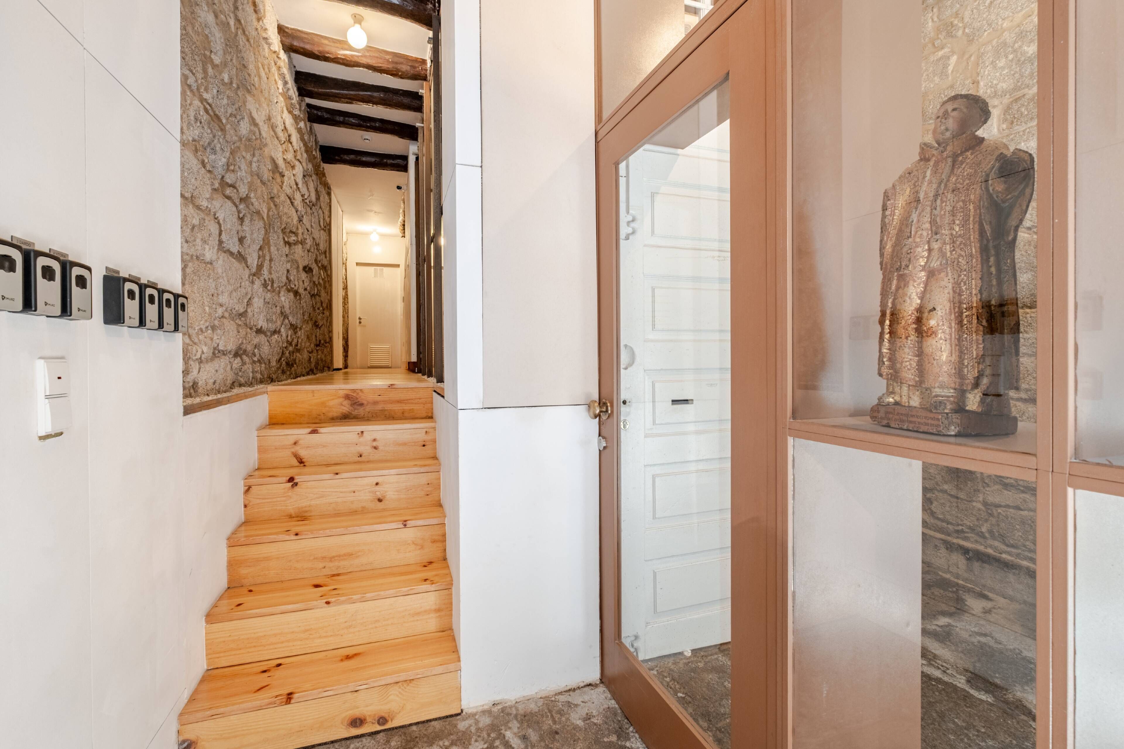 Una escalera de madera que conduce a una habitación con paredes de piedra, una puerta blanca y una estatua.