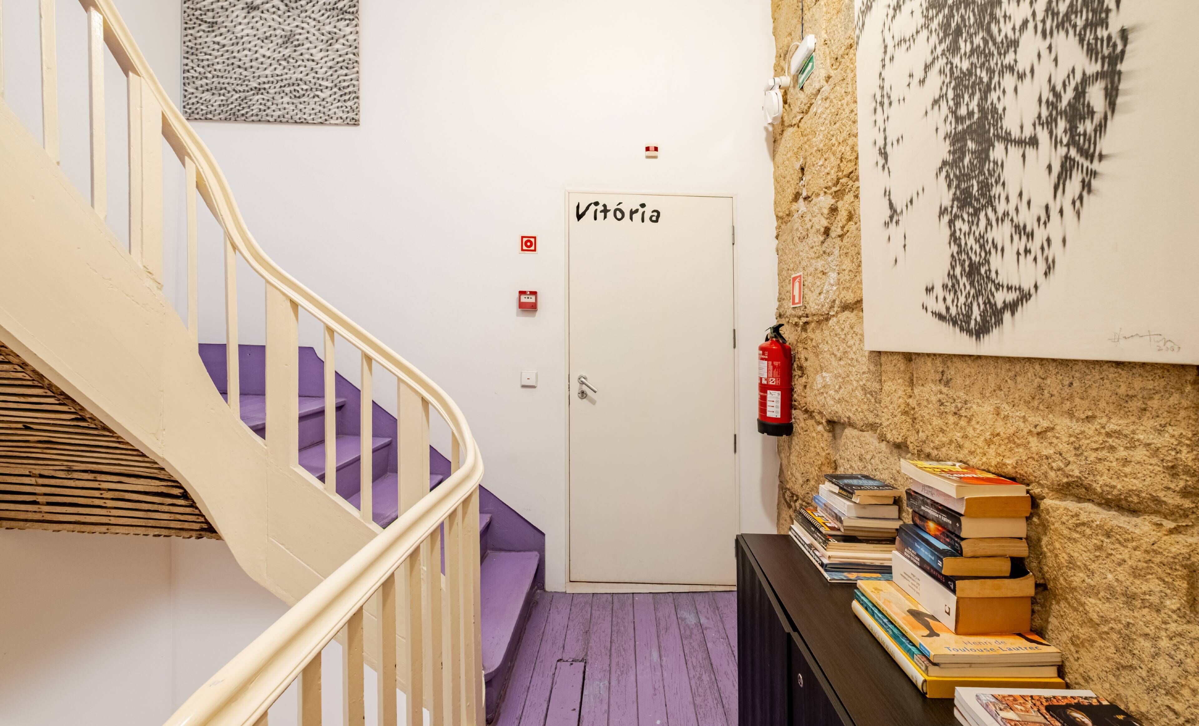 Een trap die leidt naar een kamer met de naam 'Vitória', met een brandblusser, een kunstwerk aan de muur en een stapel boeken op een tafel.