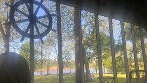 A room with large windows, a sail wheel decoration, and a view of a grassy area with trees and a body of water. - Peak Season! Arkansas River Cabin with Dock,White Bluff Views, Fishing & Sandbar (Tucker)