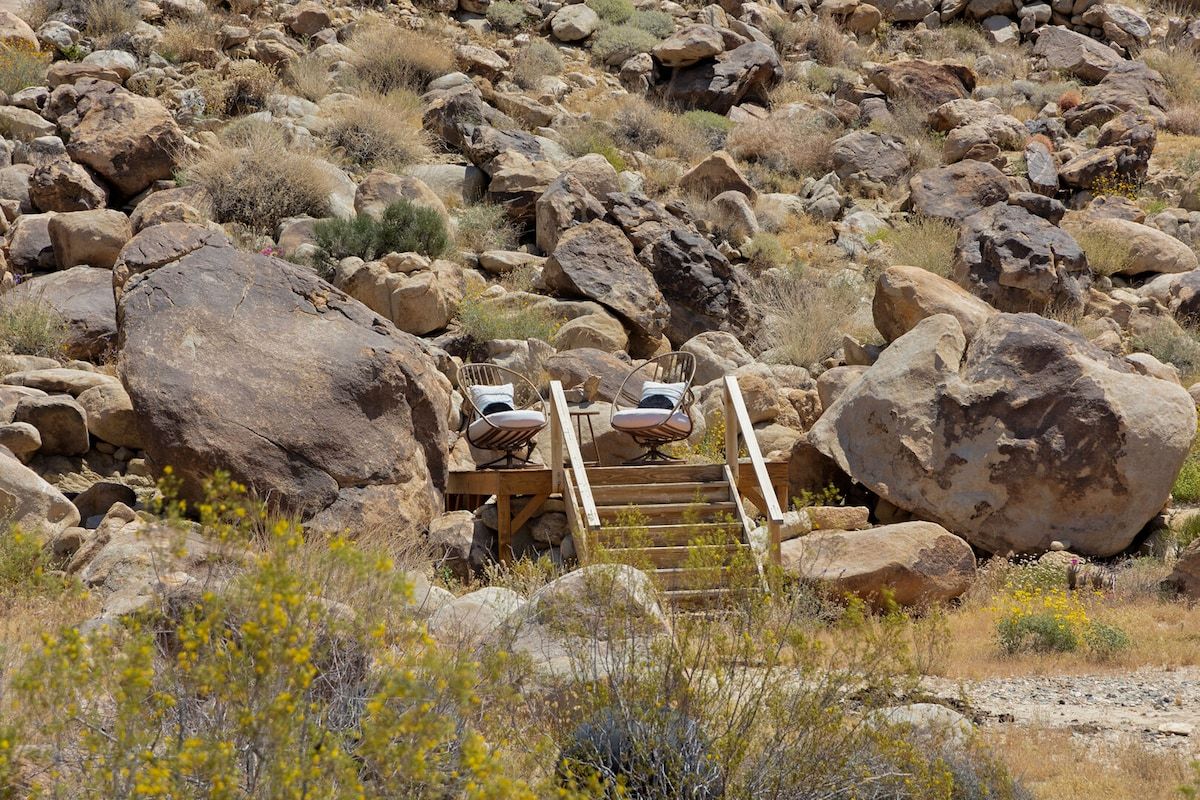 Un espace extérieur rustique, agrémenté de gros rochers, d’un escalier en bois et de chaises en métal.