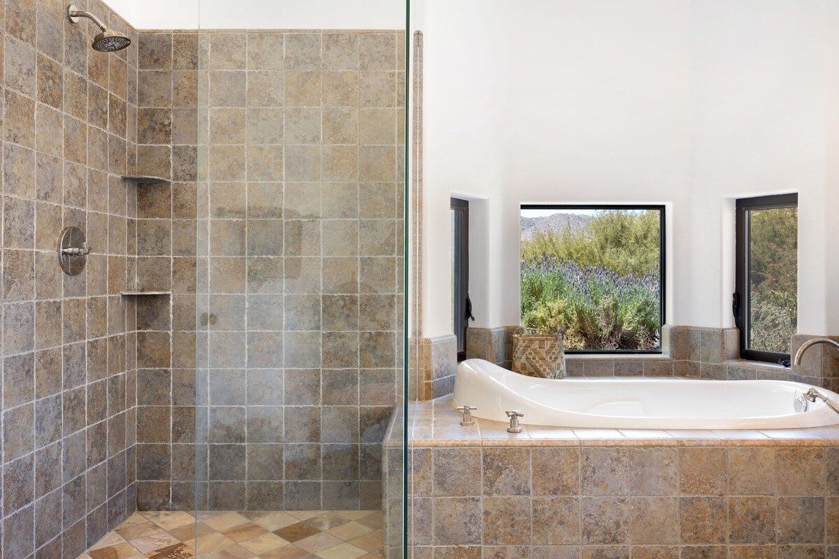 A bathroom with a shower, a bathtub, and a window with a view of greenery.