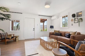 A spacious living room with a brown leather sofa, a glass coffee table, and a wicker side table. There are two windows providing natural light, and a door leading to an outdoor area. - Moonbeam Mountain - Stylish Suites + Hot Tub (Joshua Tree)