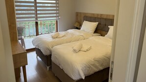 A hotel room with two beds, a wooden headboard, a desk, and a balcony with a view of greenery.