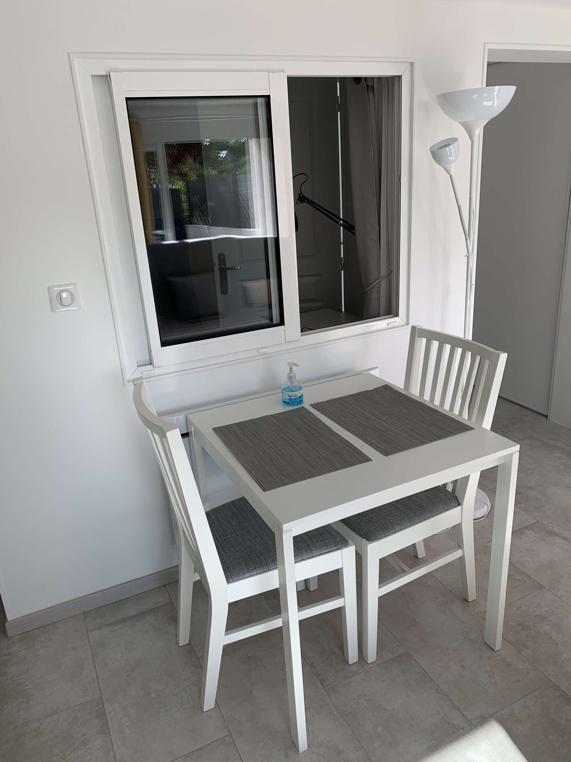 A white dining table with two chairs, a window with a view of the outside, and a floor lamp.