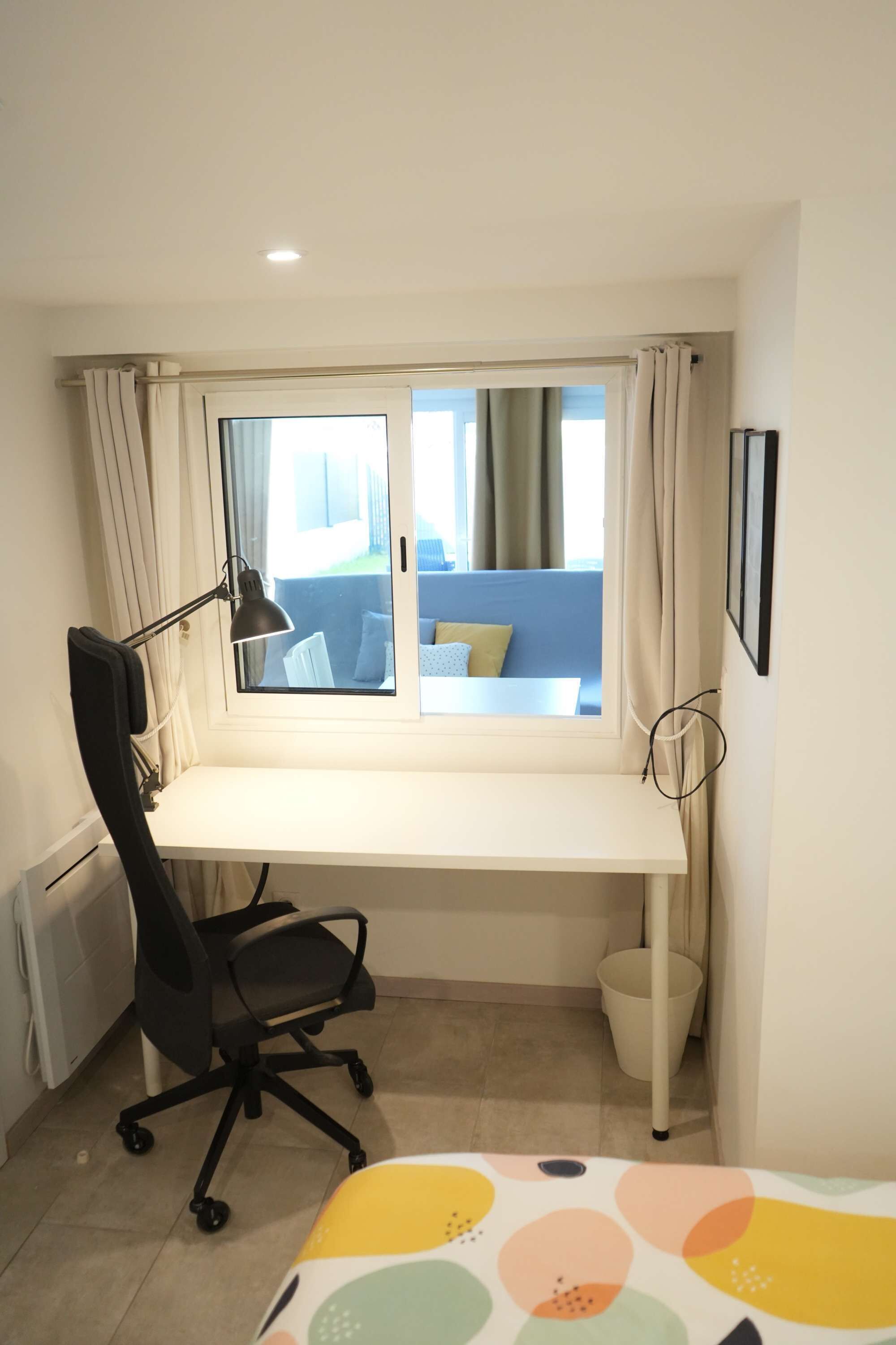 Una habitación pequeña con escritorio, silla, ventana con cortinas, cama con cabecera azul y una lámpara de pared.