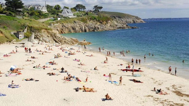 Una playa con gente tomando el sol y nadando, casas en una colina y un cielo despejado.