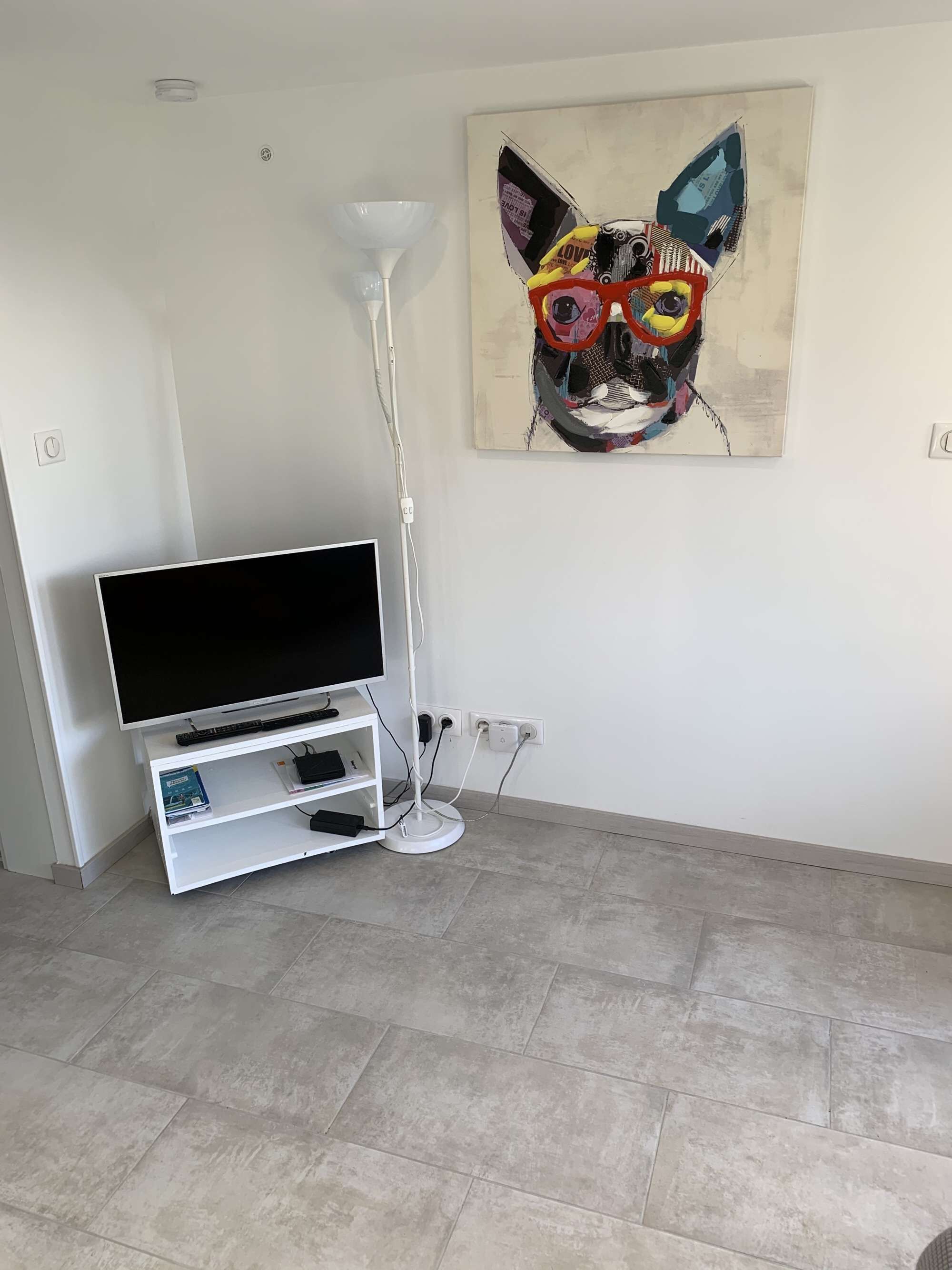 Ein modernes Wohnzimmer mit einem Flachbildfernseher auf einem weißen Schrank, einer Stehlampe und einem farbenfrohen abstrakten Gemälde an der Wand.