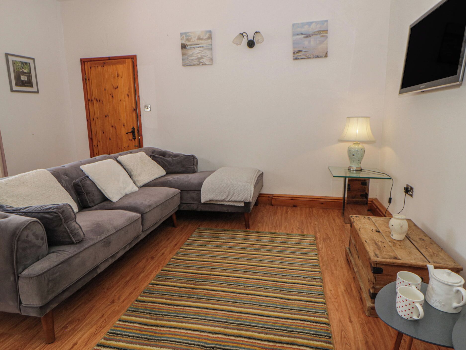 Una sala de estar con un sofá gris, una puerta de madera, un tapete a rayas, una mesita con una lámpara y un televisor montado en la pared.