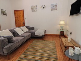 Una sala de estar con un sofá gris, una puerta de madera, un tapete a rayas, una mesita con una lámpara y un televisor montado en la pared.