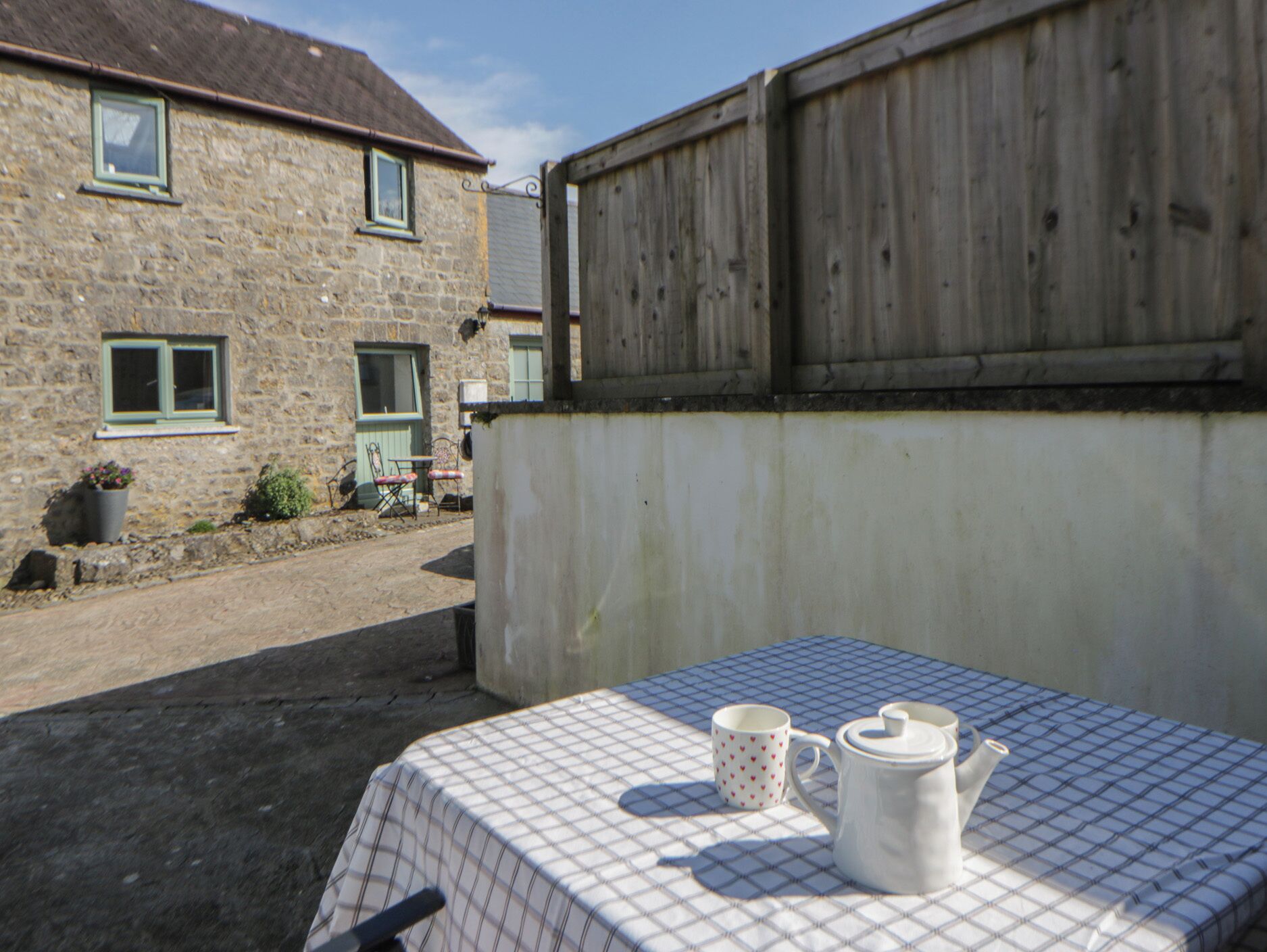Una mesa con mantel de cuadros, una taza blanca y una jarra blanca en un patio soleado con edificios de piedra al fondo.