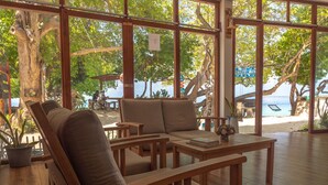 A wooden lounge area with seating and a table, large windows offering a view of the beach and trees. - Thundi village and spa (Maalhos)