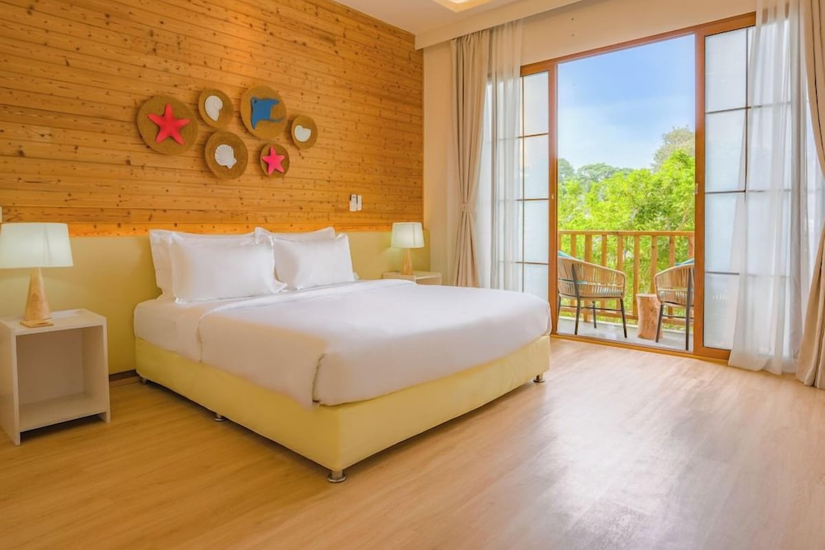 Quarto de hotel com cama grande, painéis de parede de madeira, varanda com mobiliário exterior e vista para o verde.