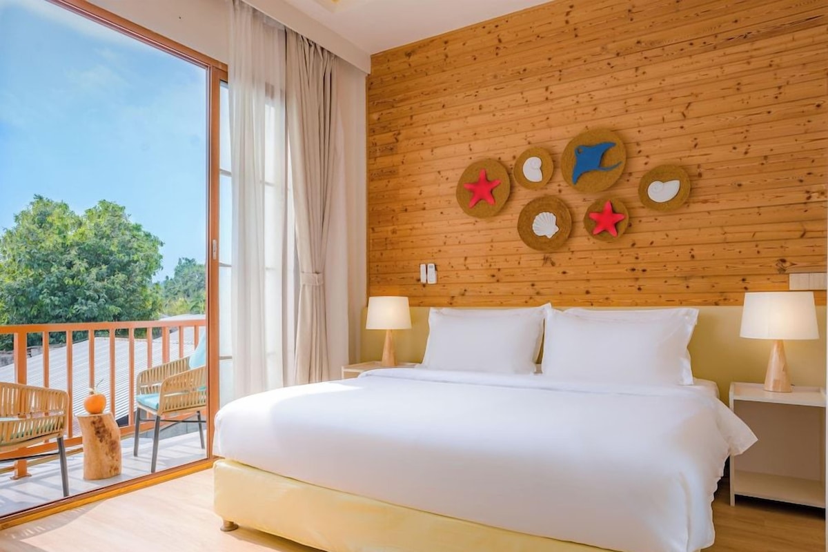 Quarto de hotel com uma cama grande, duas mesas de cabeceira com luminárias, uma parede de madeira com objetos decorativos, uma varanda com cadeiras de vime e vista para árvores.