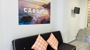 Een modern interieur met een zwarte bank, twee kussens met oranje ruitjespatroon, een lijst met een foto van een zonsondergang over de zee, en een aan de muur bevestigde televisie.