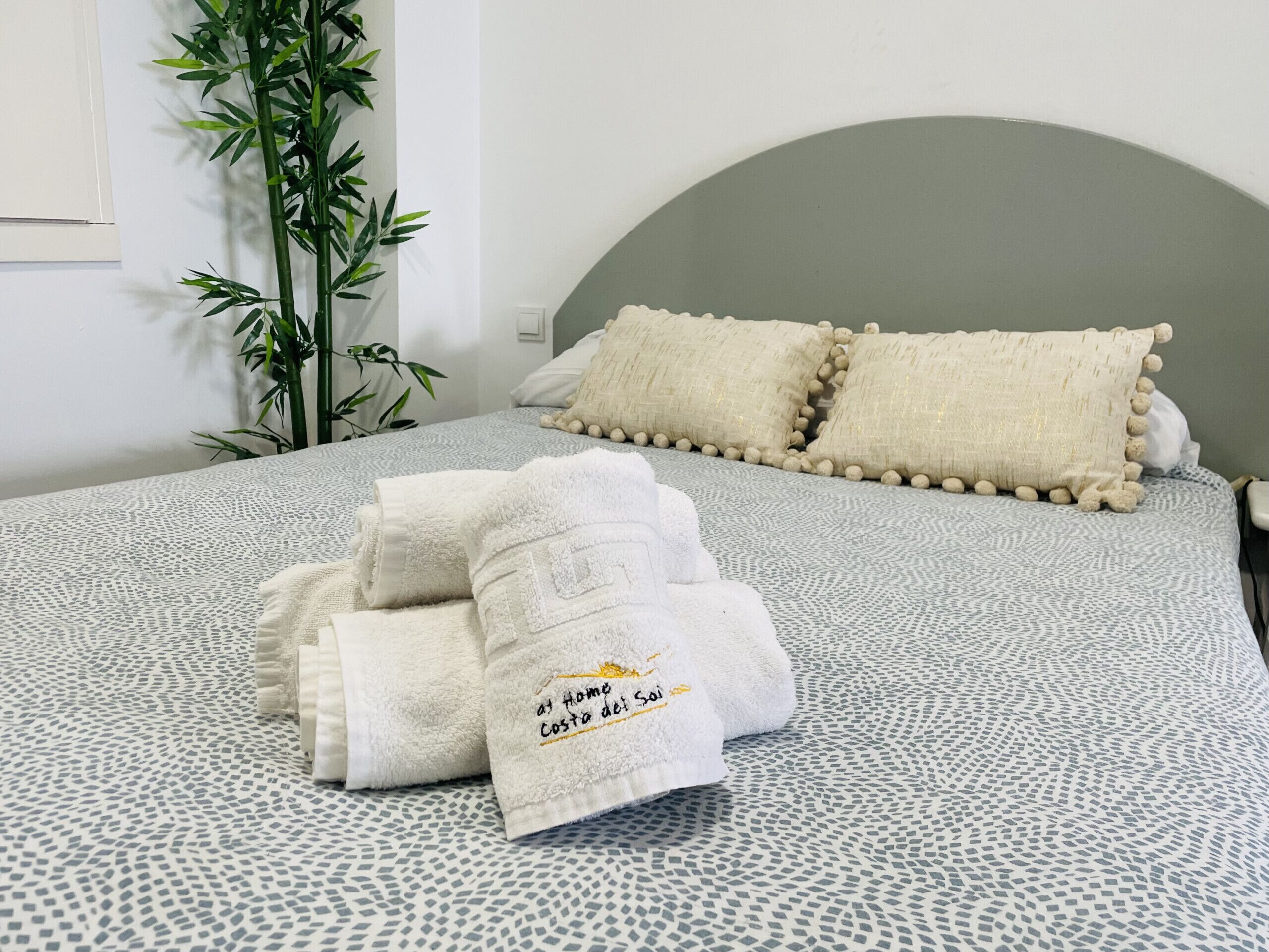 A neatly made bed with a patterned bedspread, two pillows, and a stack of folded towels with a logo.
