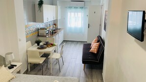 Een compacte leefruimte met een kitchenette, een eethoek, een bank en een aan de muur bevestigde televisie.