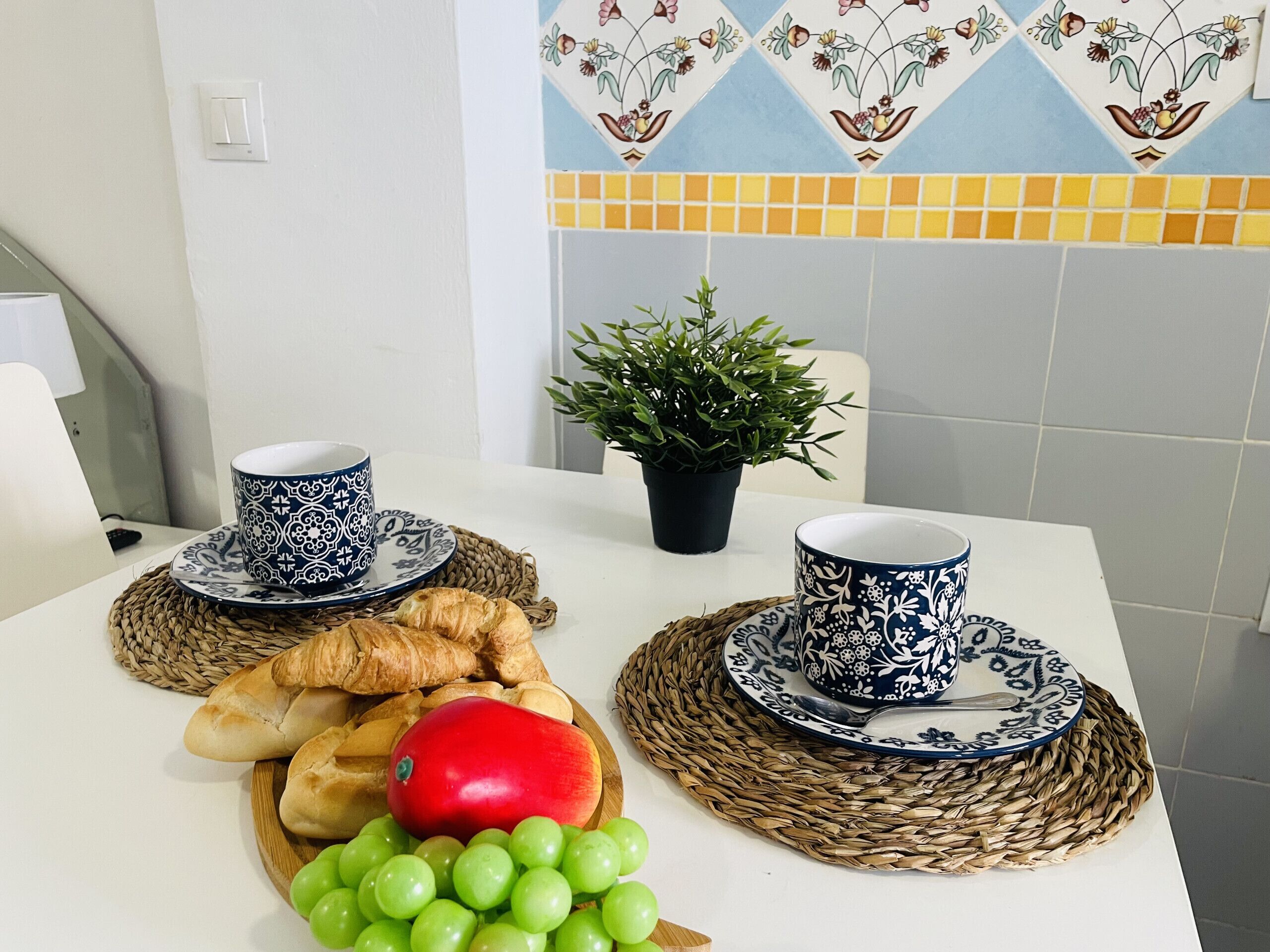 A breakfast setting with two cups, a croissant, a red apple, and green grapes on a table.
