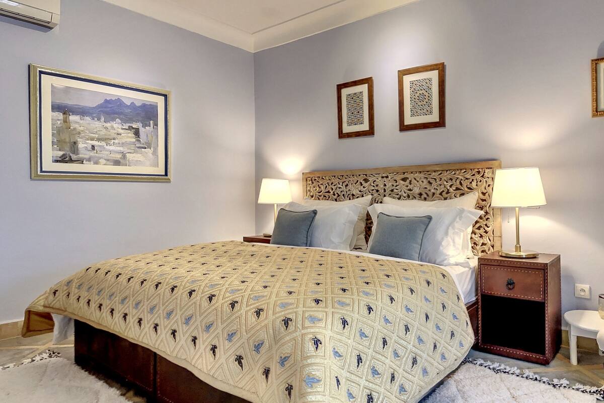 A neatly arranged bedroom with a large bed, two bedside tables with lamps, a decorative headboard, and framed artwork on the walls.
