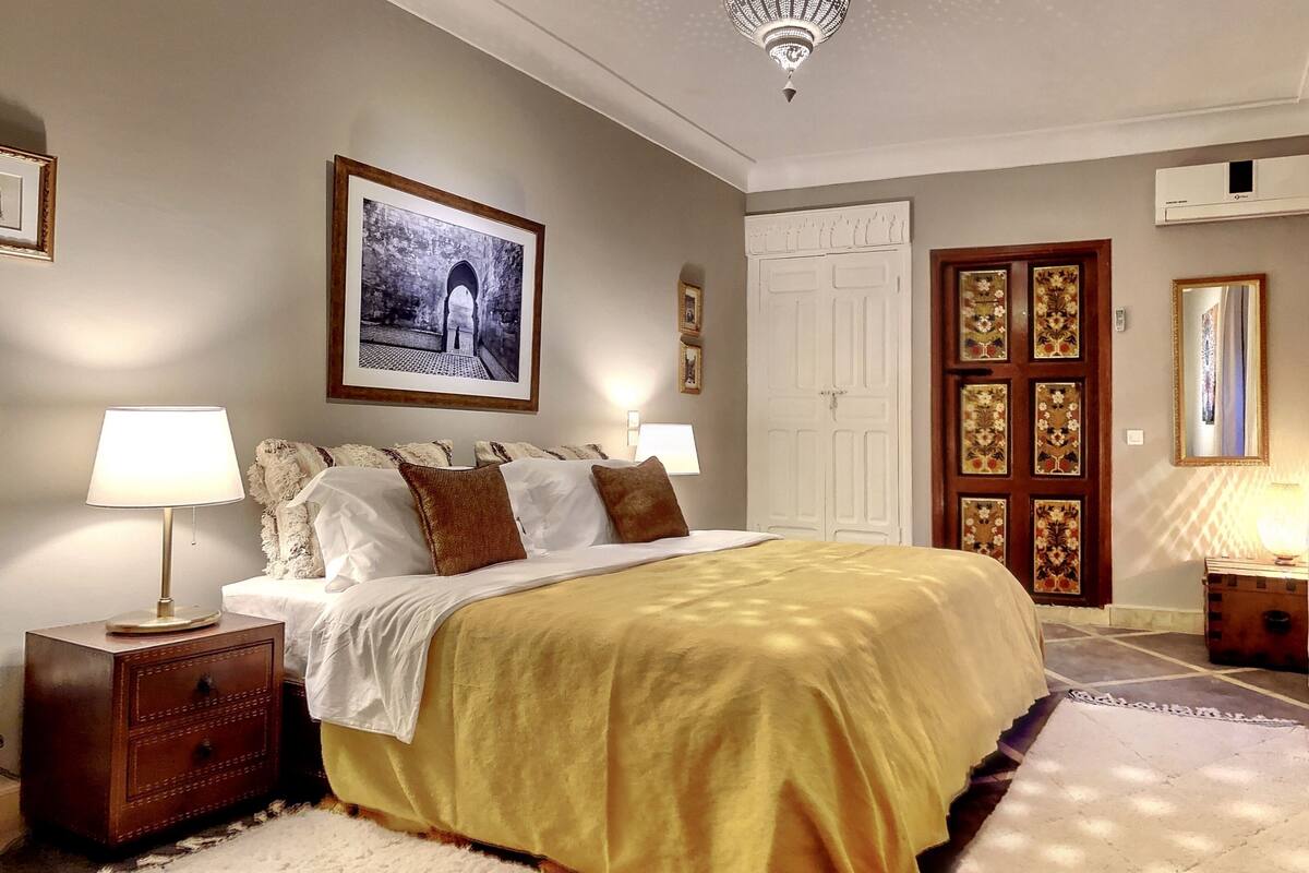 A bedroom with a bed, two bedside tables, a nightstand, a large framed picture, a door with decorative panels, and a mirror.