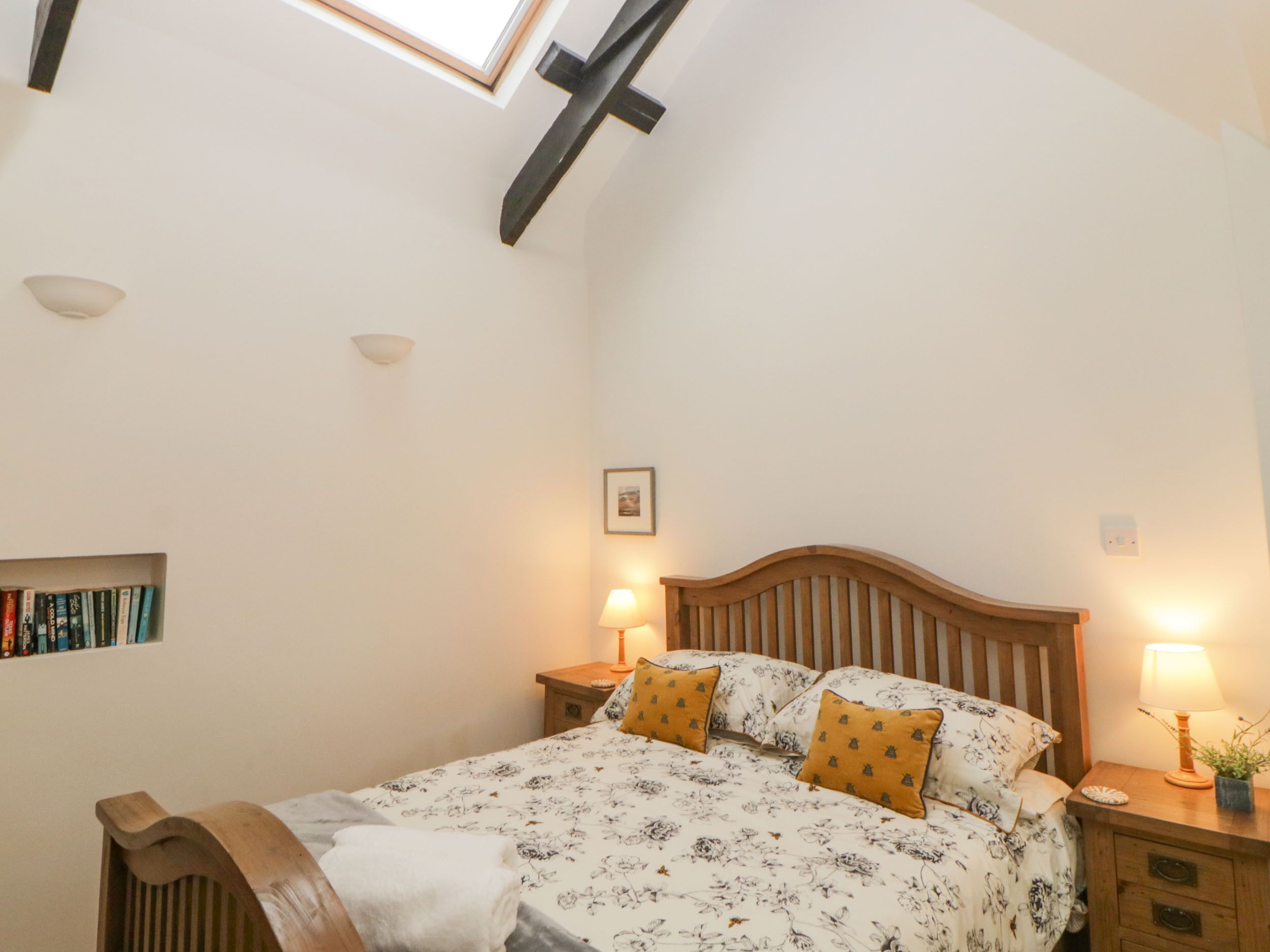 A bedroom with a sloped ceiling, a bed with floral bedding, two bedside lamps, and a small bedside table with a plant.