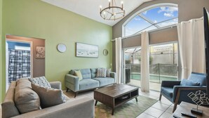 A living room with a sofa, a coffee table, and a TV stand. There is a window with curtains and a view of the outside.