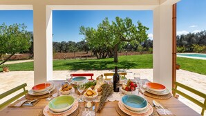 Un área de comedor al aire libre con una mesa lista para una comida, rodeada de sillas y con vista a un jardín ajardinado con árboles y una piscina.