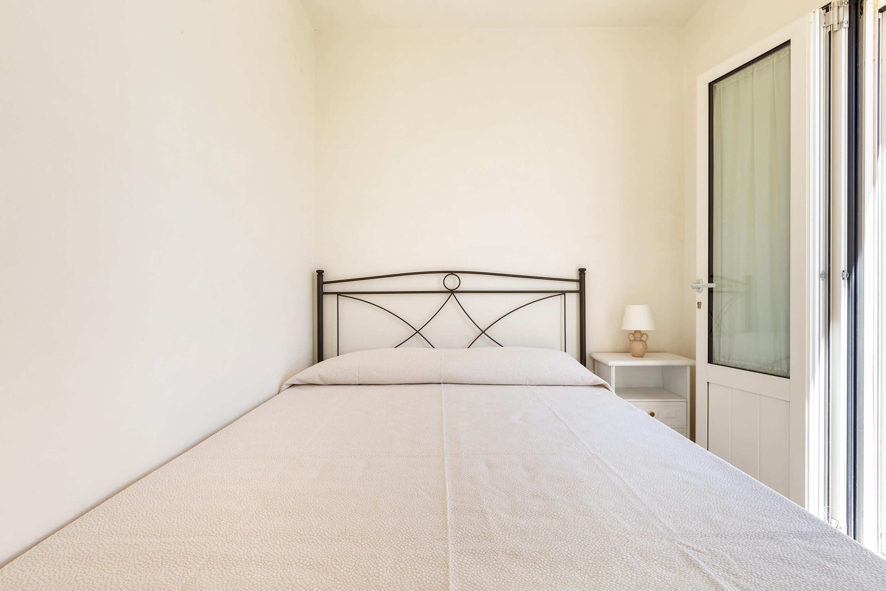 Una cama individual con estructura metálica y cabecera sencilla, una mesita de noche con lámpara, y una puerta con panel de vidrio satinado.