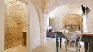 Una habitación con pared de piedra, una cama con cabecera de madera, una mesa con mantel de encaje y una silla.