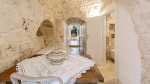 Una habitación con paredes de piedra, una mesa de madera, un mantel blanco y una silla de mimbre.