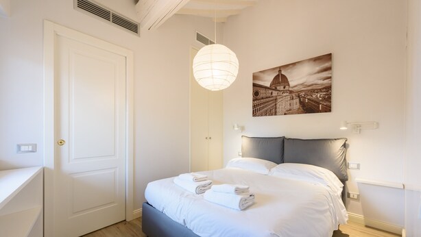 A bedroom with a bed, a white door, a hanging light fixture, and a cityscape painting on the wall. - Santa Croce Top by Mmega (Florence)