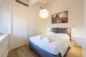 Un dormitorio con cama, una puerta blanca, una lámpara colgante y un cuadro con una vista urbana en la pared.