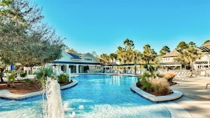 A large outdoor swimming pool with a central fountain, surrounded by palm trees and a building with a sloped roof.