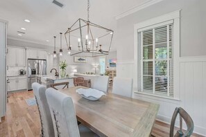 Una cucina moderna con mobili bianchi, frigorifero in acciaio inossidabile, illuminazione a sospensione e zona pranzo con tavolo e sedie in legno.