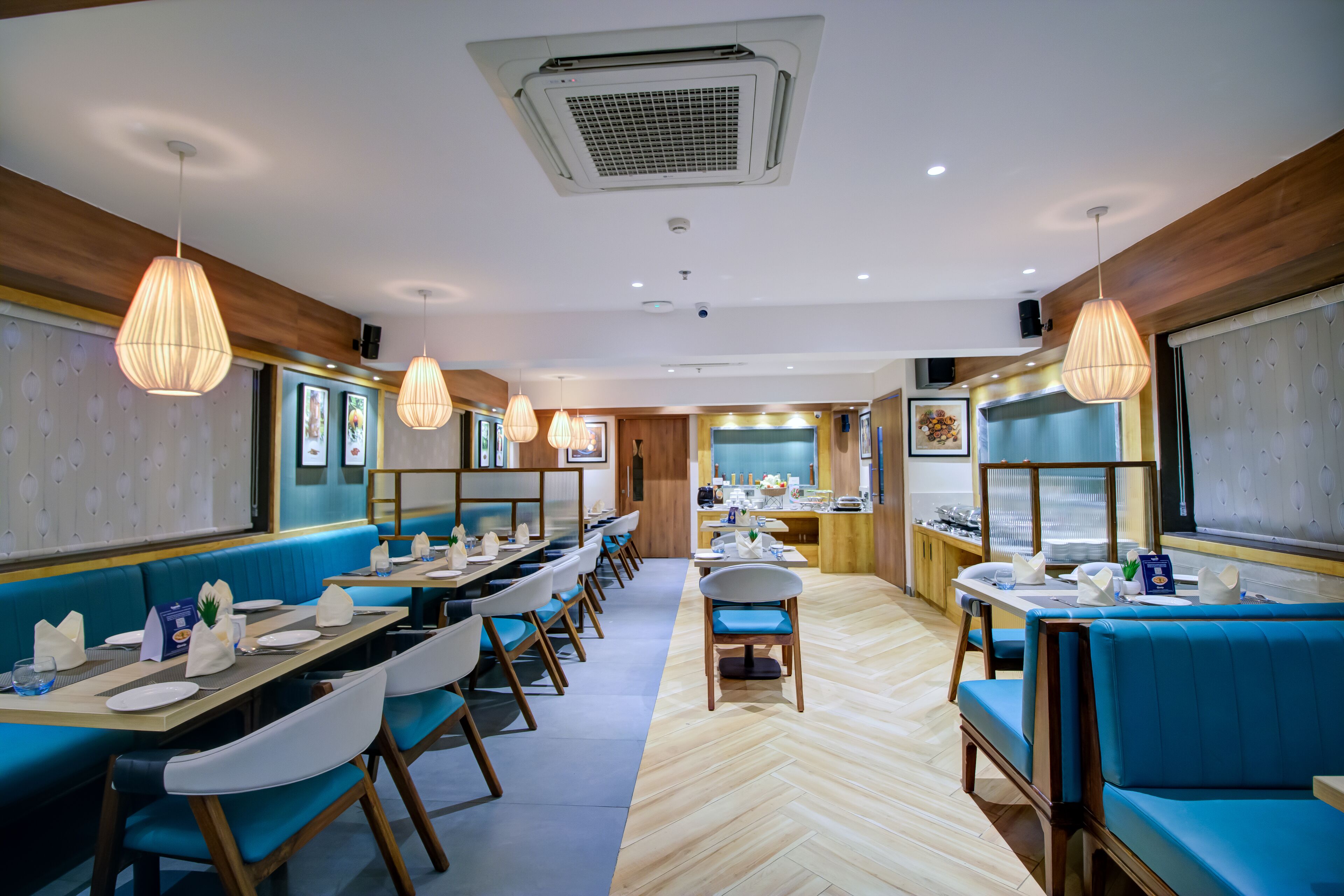 Un restaurante con largas mesas compartidas, asientos tipo banca acolchados en azul, lámparas colgantes y un patrón de suelo en espiga.