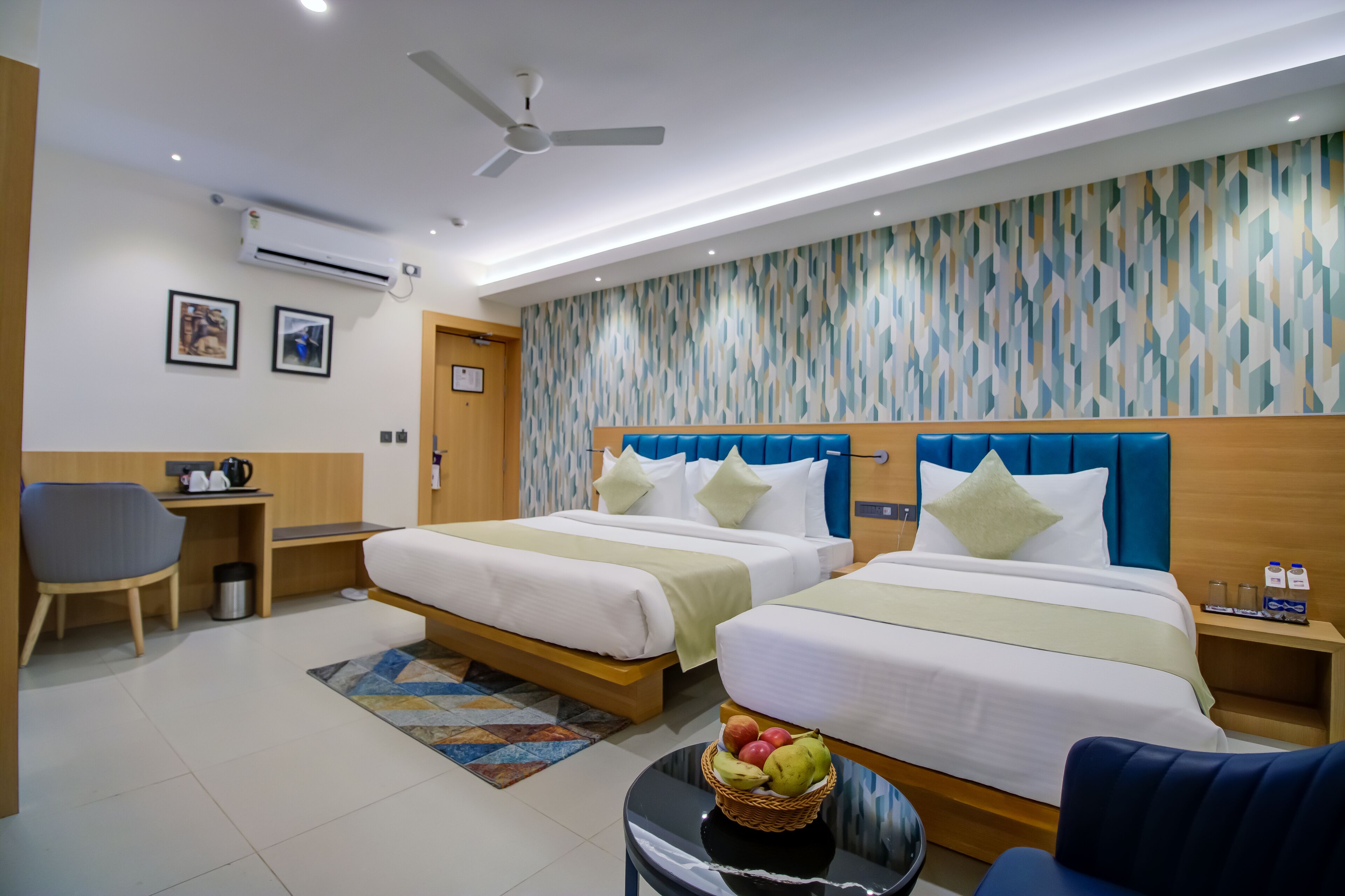 A hotel room with two beds, a desk, a chair, and a small table with fruit.