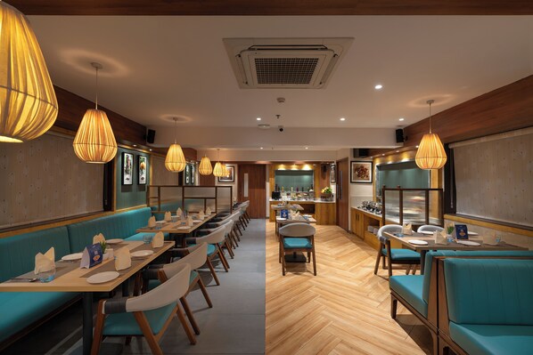 Un restaurante con largas mesas compartidas, asientos tipo banca acolchados en azul y lámparas colgantes.