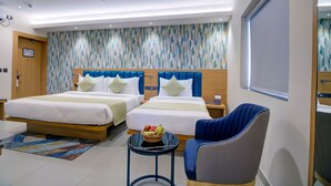 Una habitación de hotel moderna con dos camas, un ventilador de techo, una mesita con fruta y una silla azul.