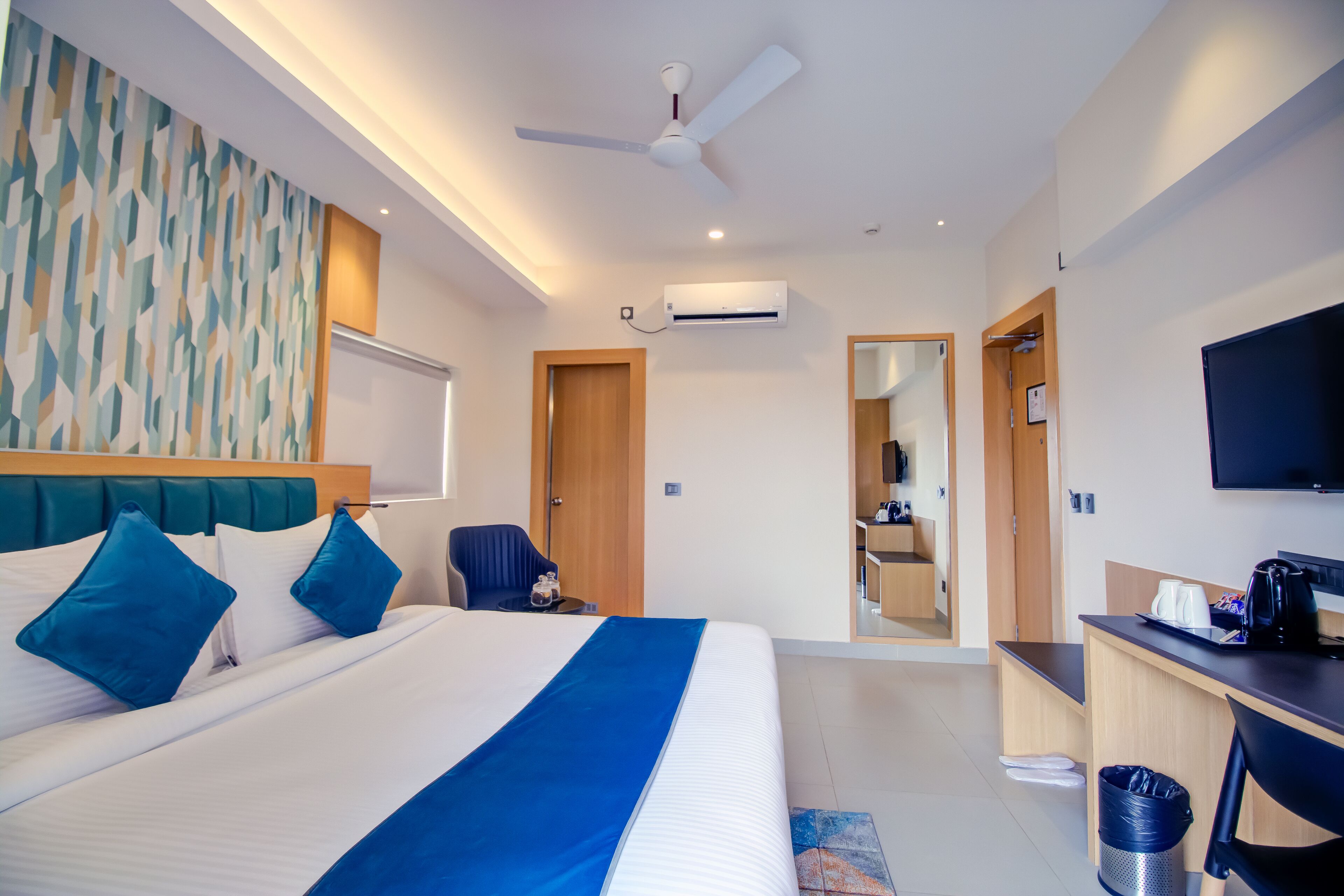 A modern hotel room with a large bed, a desk, a chair, a TV, and an air conditioning unit.