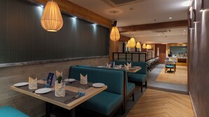 Un restaurante con asientos tipo banca en color verde azulado, mesas de madera e iluminación colgante.