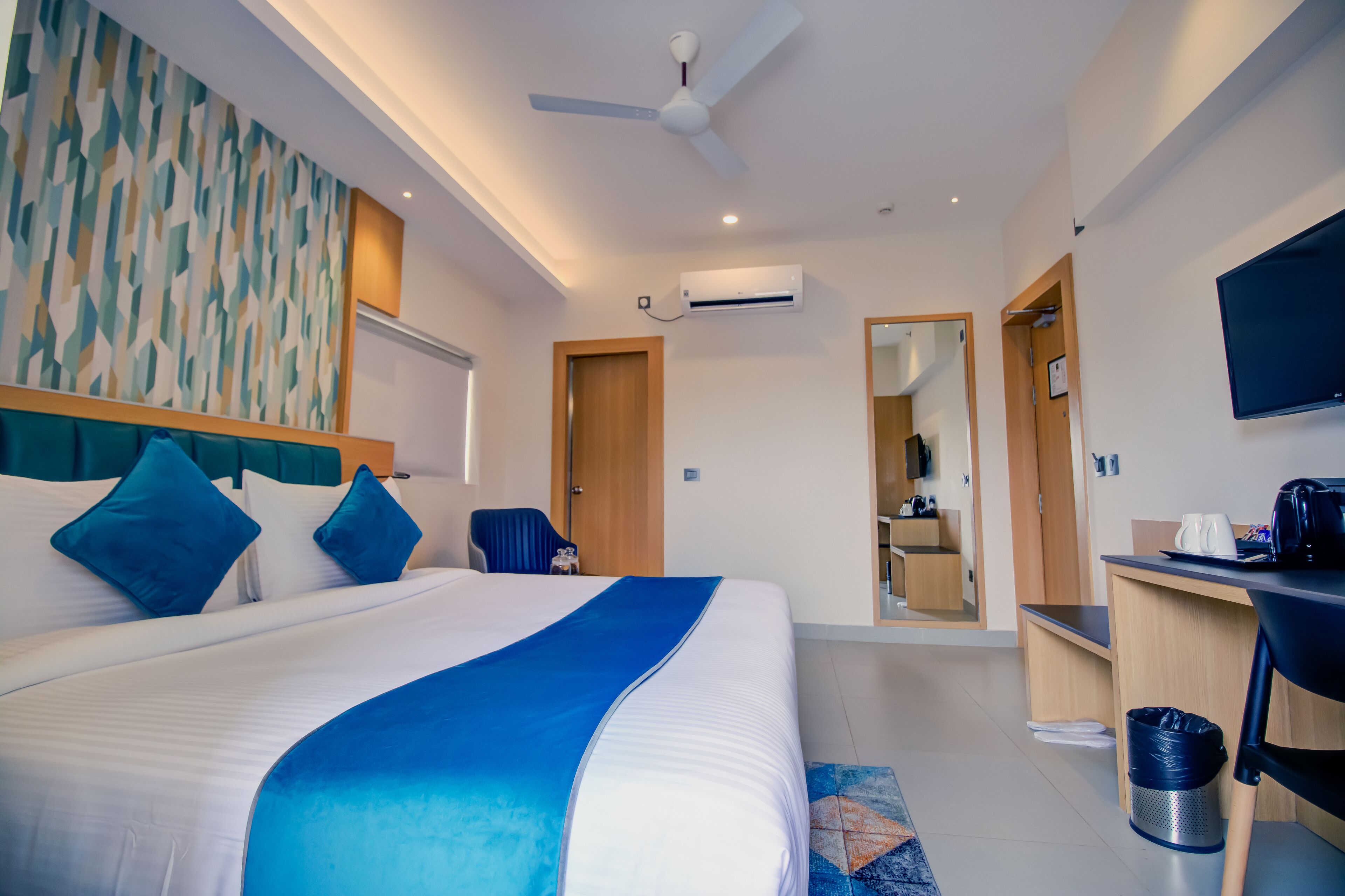A modern hotel room with a large bed, a ceiling fan, an air conditioning unit, a desk, a television, and a mirror.