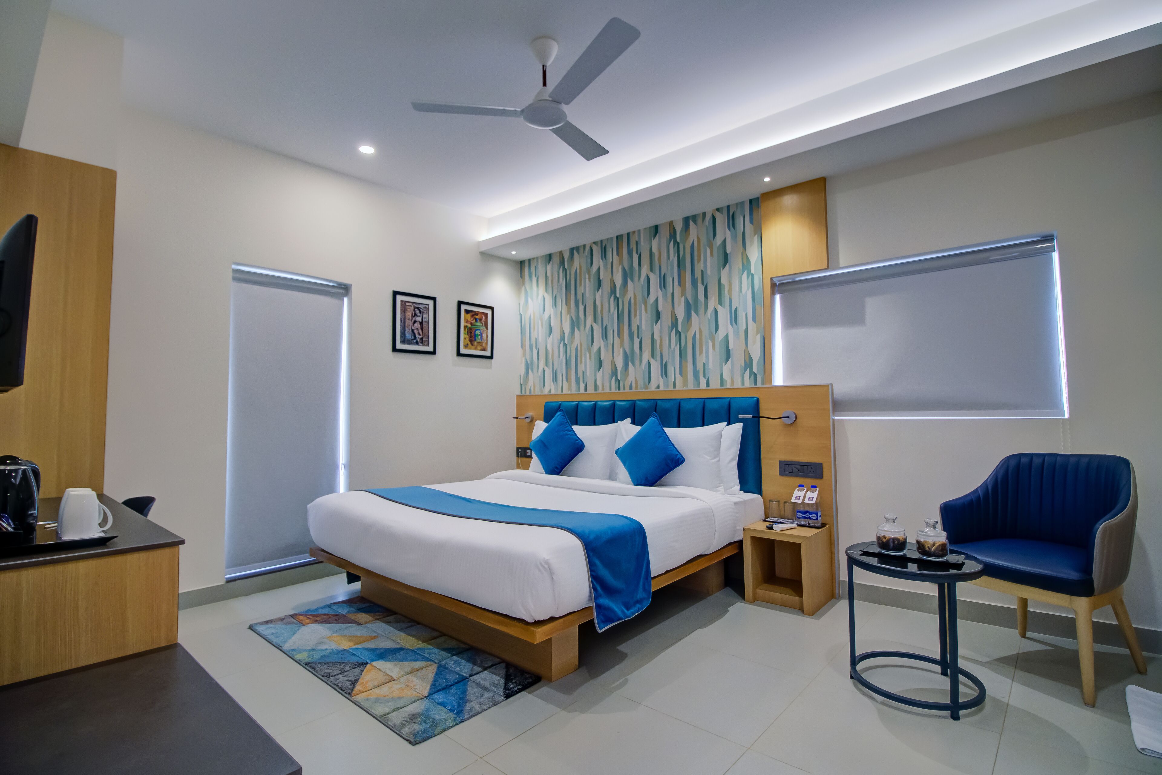 A modern hotel room with a large bed, a desk with a coffee maker, a chair, a small table with drinks, and a ceiling fan.