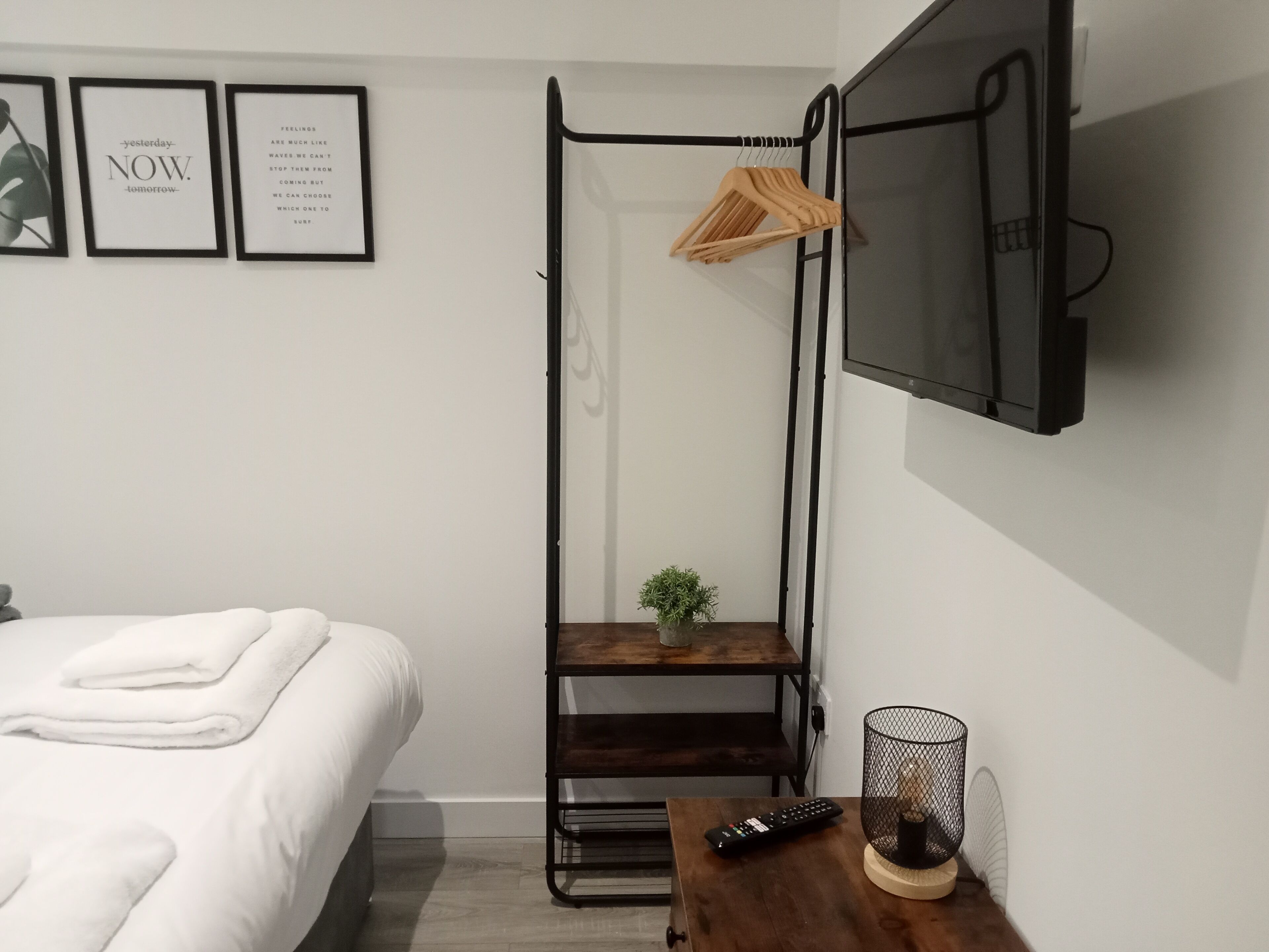 Ein Schlafzimmer mit einem Bett, einem an der Wand montierten Fernseher, einer kleinen Ablage mit einer Pflanze und einem Holztisch mit Fernbedienung.