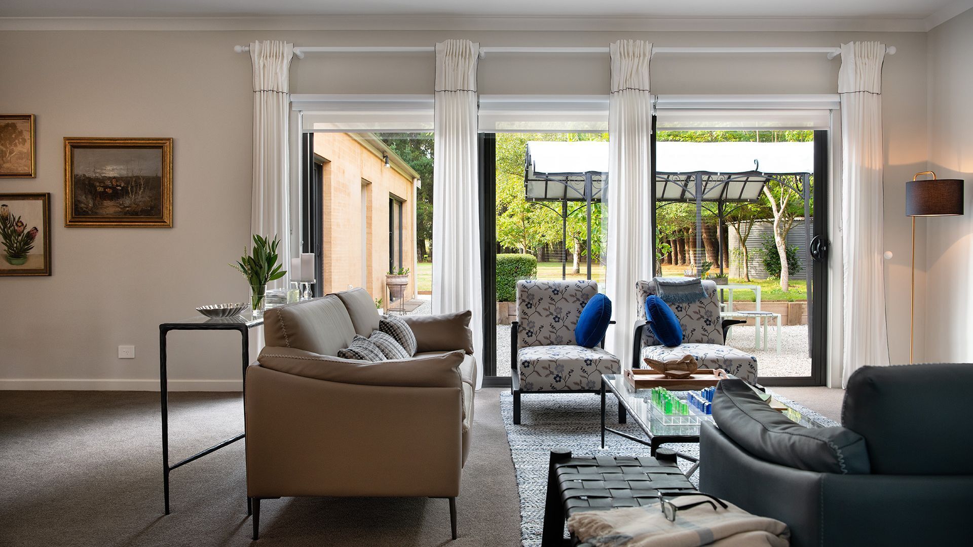 Een woonkamer met een beige bank, een fauteuil met patroon en een donkere fauteuil. Er staat een salontafel met een dienblad en glazen op. De kamer heeft grote ramen met witte gordijnen en uitzicht op een tuin en een terras.