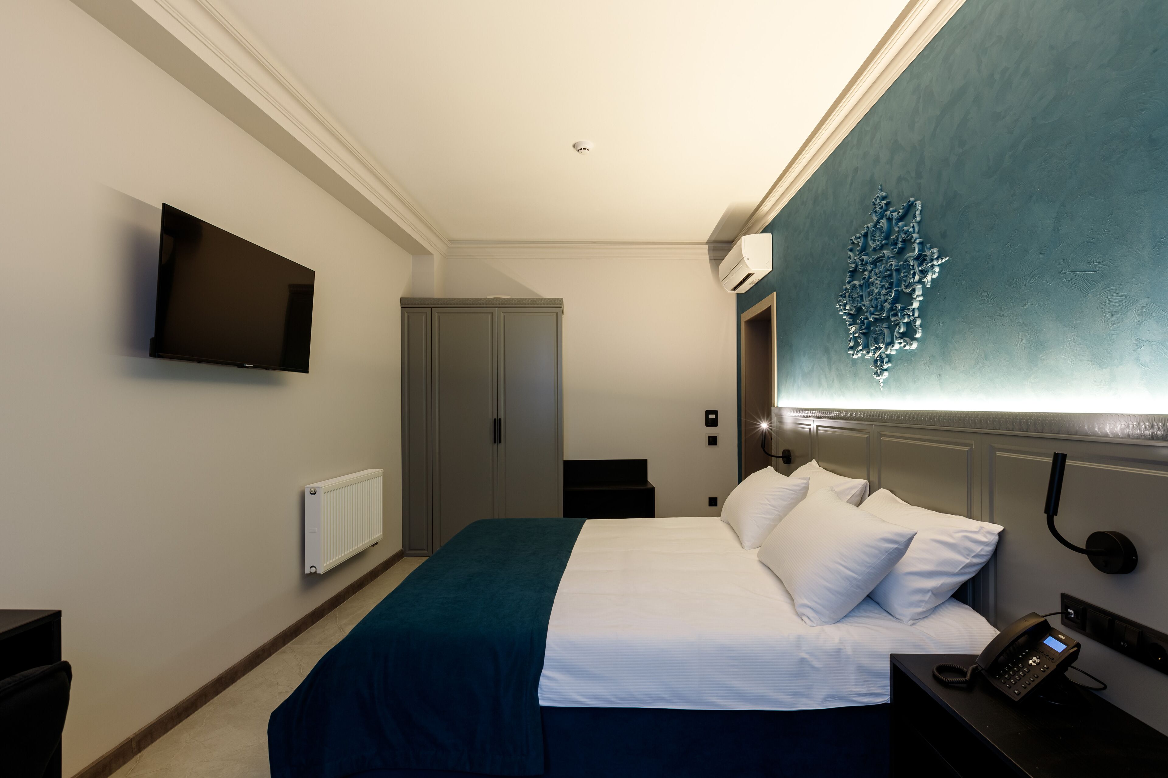 Una habitación de hotel con cama, televisor de pantalla plana montado en la pared, teléfono junto a la cama y aire acondicionado también en la pared.