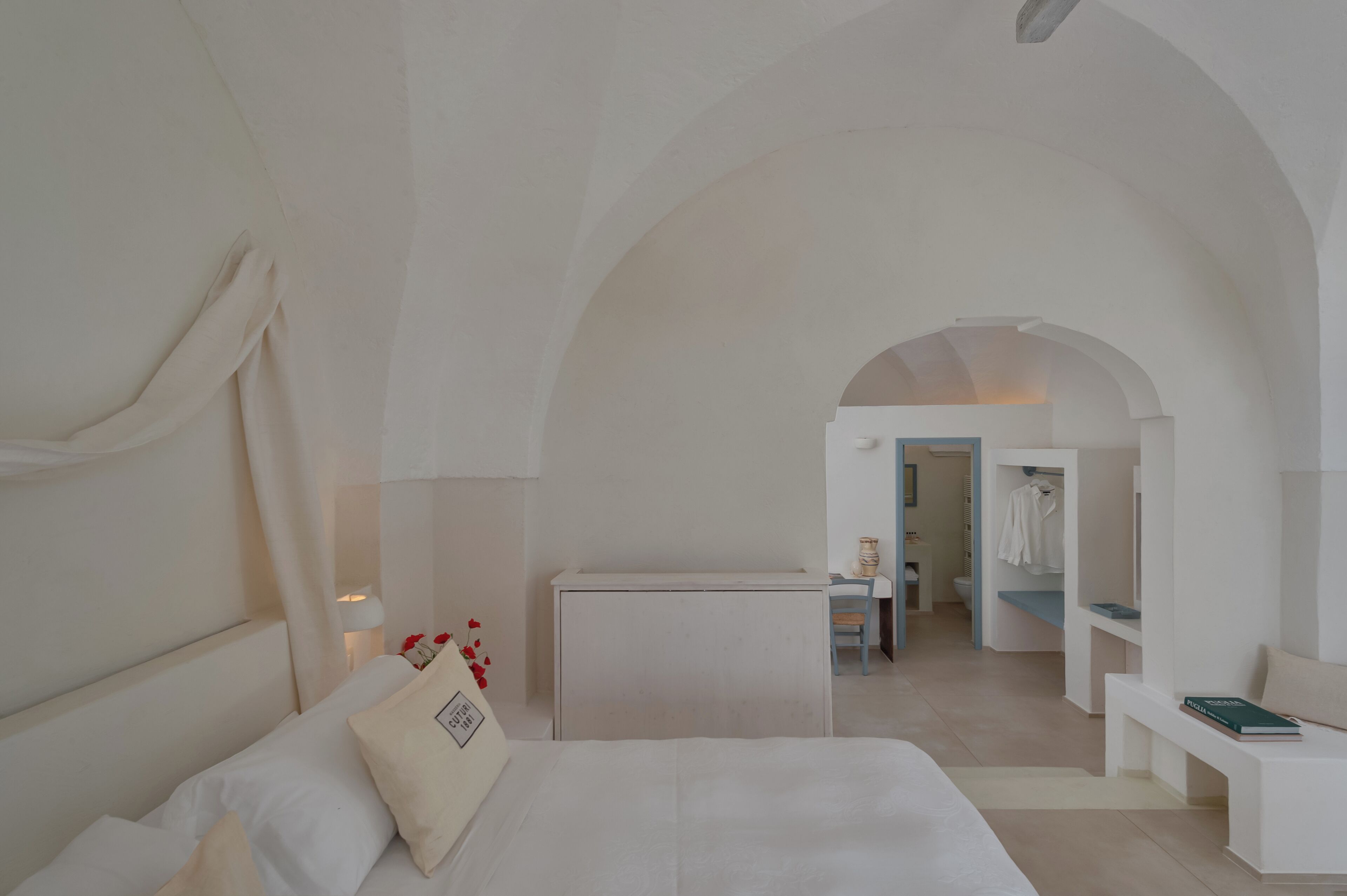 A minimalist bedroom with a large bed, a desk, and an arched doorway leading to a bathroom.