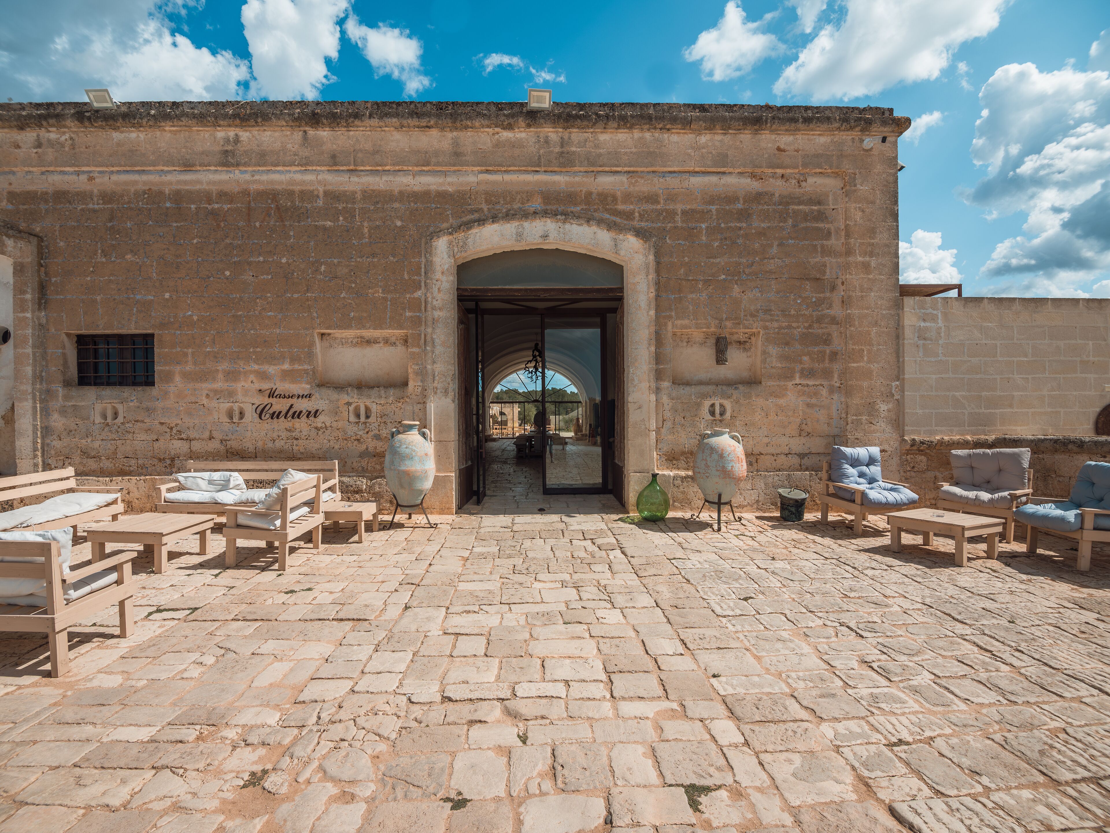 A courtyard with stone flooring, a central archway, and seating areas with cushions and tables.