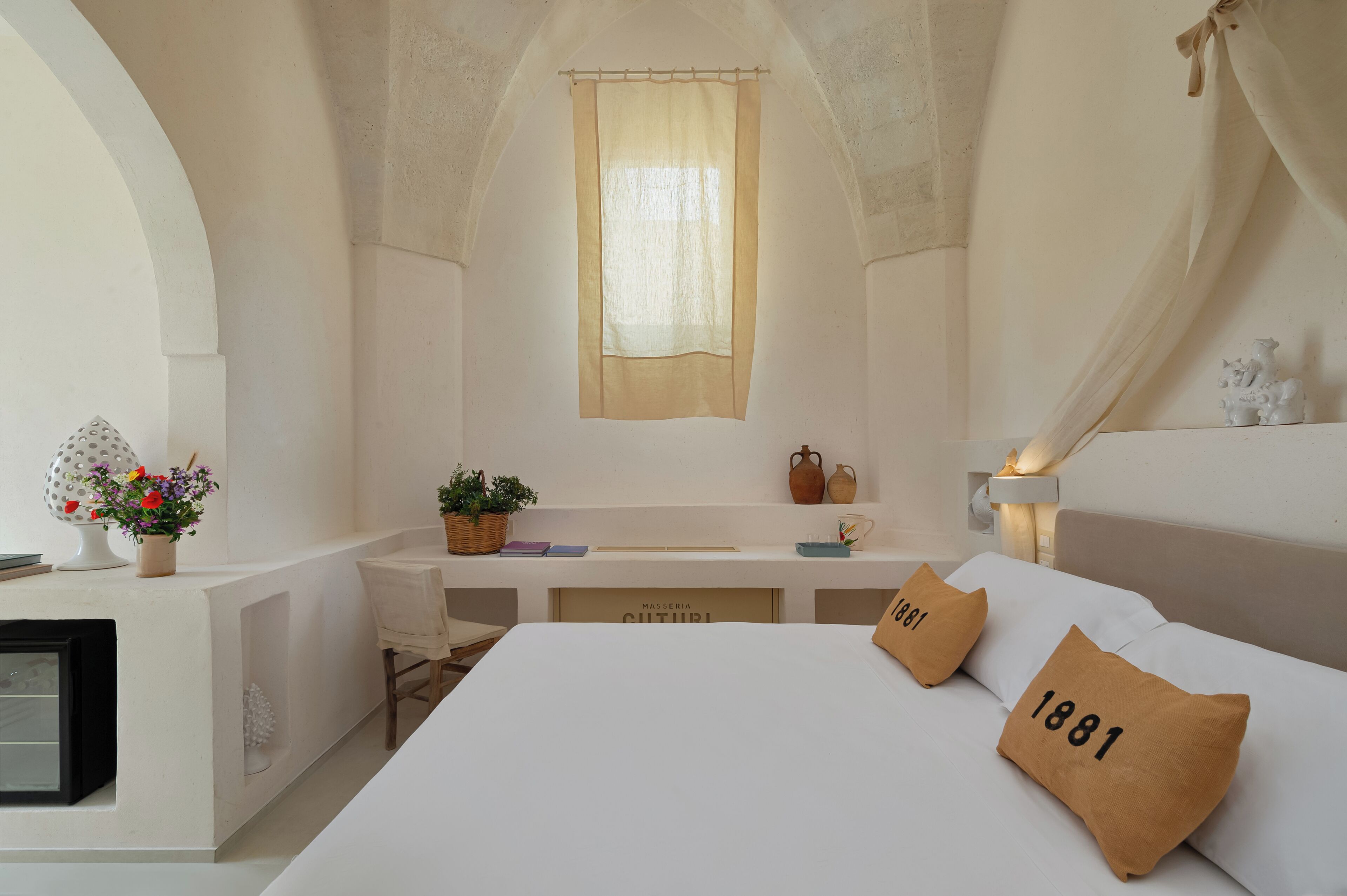 Un dormitorio con cama, mesita de noche, ventana con cortina translúcida y una pequeña mesa con lámpara.