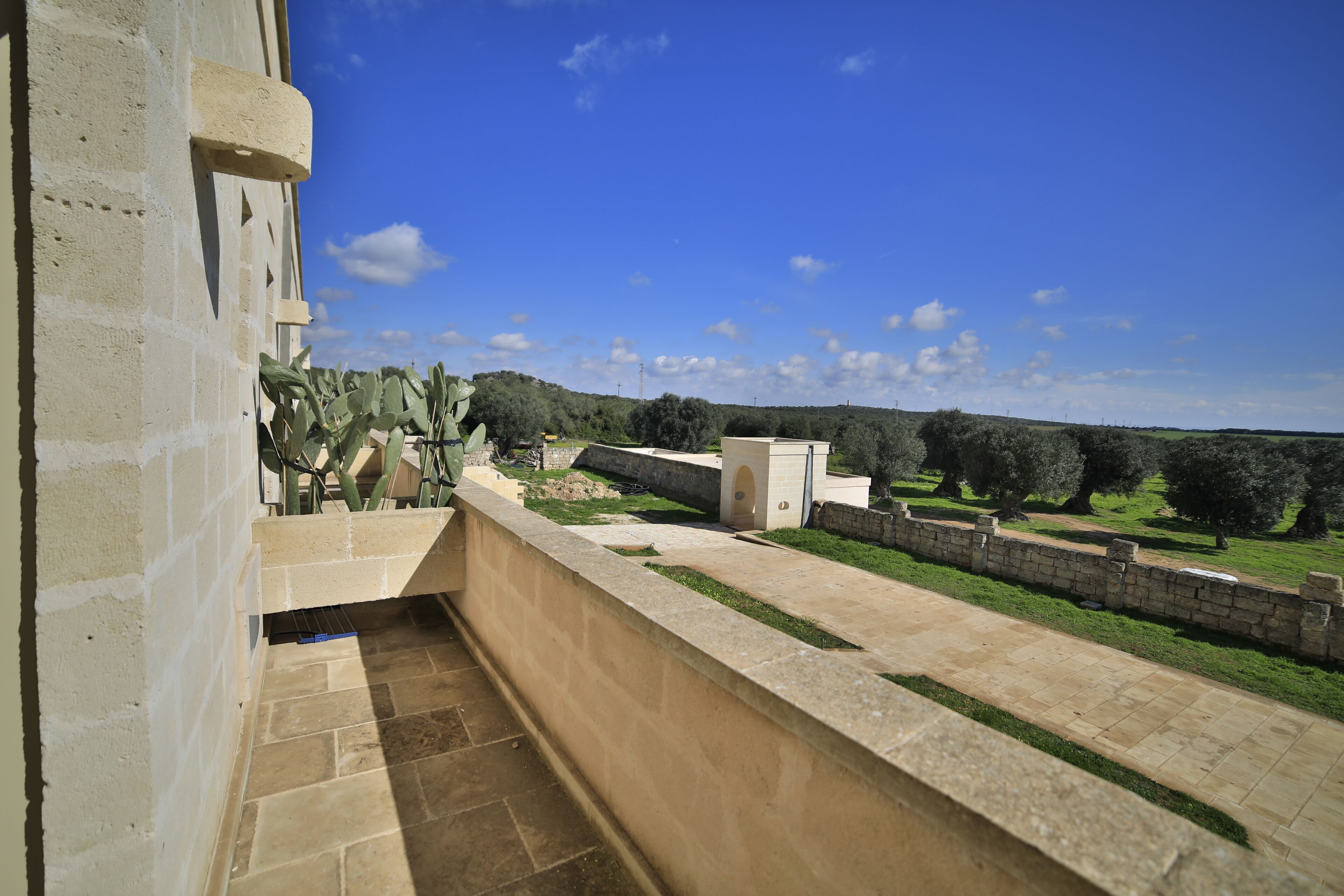 Un balcon donnant sur un jardin aménagé, un mur de pierre et un ciel d’un bleu limpide.
